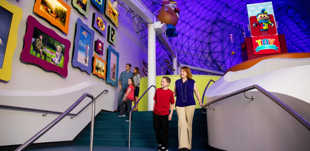 Two families walking down the stairs at The Strong Museum while chatting about the different images of Play that are hung on the wall.